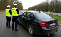 zdjęcie kolorowe na którym widać nieznakowany czarny radiowóz m-ki BMW i policjantów dwóch którzy patrzą i obserwują sytuację na drodze. W tle widać drogę i jadące pojazdy.