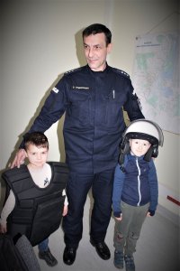zdjęcie kolorowe na którym stoi policjant  w mundurze  z dwoma przedszkolakami ubranymi w kask i kamizelkę policyjną