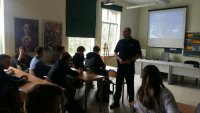 zdjęcie na którym widać jak policjant przeprowadza prelekcje w klasie z uczniami i wyświetlona jest w tle prezentacja multimedialna