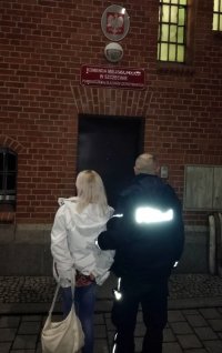 Fotografia kolorowa. Umundurowany policjant stoi wraz z zatrzymaną poszukiwaną kobietą przez wejściem do pomieszczenia dla osób zatrzymanych.