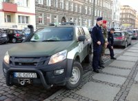 Zdjęcie kolorowe. Na zdjęciu zaparkowany radiowóz Żandarmerii Wojskowej. Obok stają policjanci i funkcjonariusze Żandarmerii Wojskowej i obserwują miejsce zagrożone. W tle widać ulice, pojazdy zaparkowane na wyznaczonych miejscach oraz kamienice.