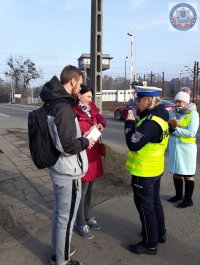 Fotografia kolorowa. Na zdjęciu policjantka ubrana w mundur i kamizelkę odblaskową rozmawia z przechodniami wręczając im ulotki informacyjne. W tle widać przejazd kolejowy