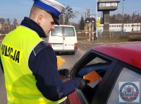 Policjant Wydziału Ruchu Drogowego ubrany w mundur i kamizelkę odblaskową z napisem Policja wręcza kierującemu ulotki informacyjne, który jedzie samochodem koloru czerwonego. W tle widać  busa koloru białego przejeżdżającego przez przejazd kolejowy z zaporami