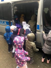 zdjęcie kolorowe które przedstawia radiowóz policyjny bus, z otwartymi bocznymi dzrzwiami. W samochodzie siedzą dzieci a część z nich stoi przed radiowozem. ubrane są w kolorowe kurtki.